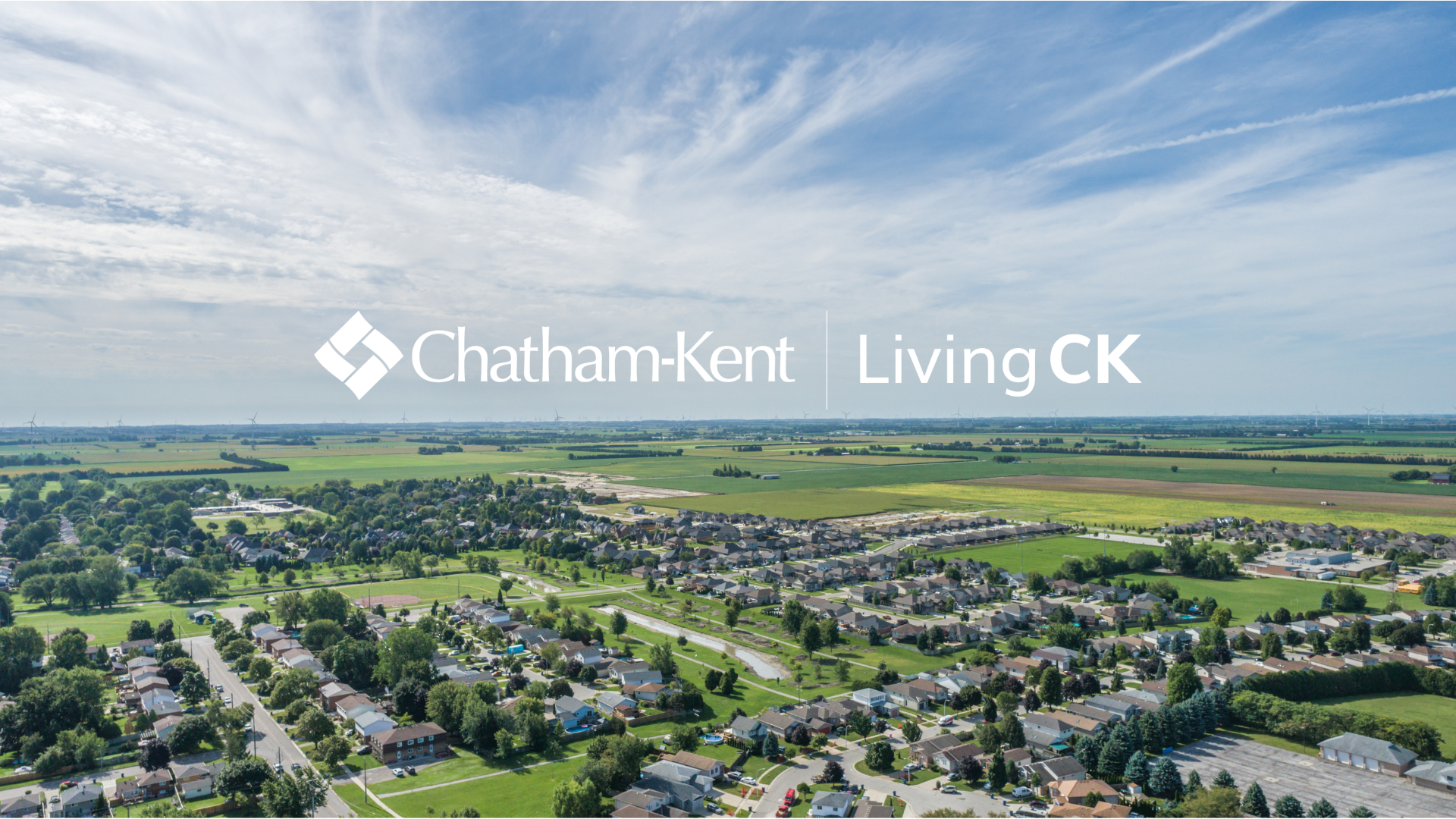 Chatham-Kent drone shot of neighbourhoods with a creek running through the middle and green fields in the background. The Municipality of Chatham-Kent LivingCK logo is overlaid in the center in white.